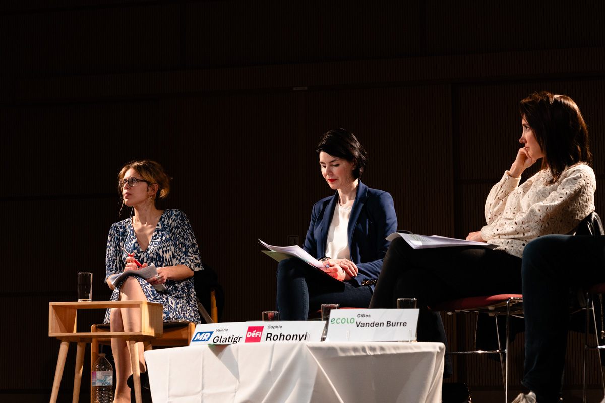 Three panellists seated in side light on a dark stage, papers on their laps, one looking down at her notes.