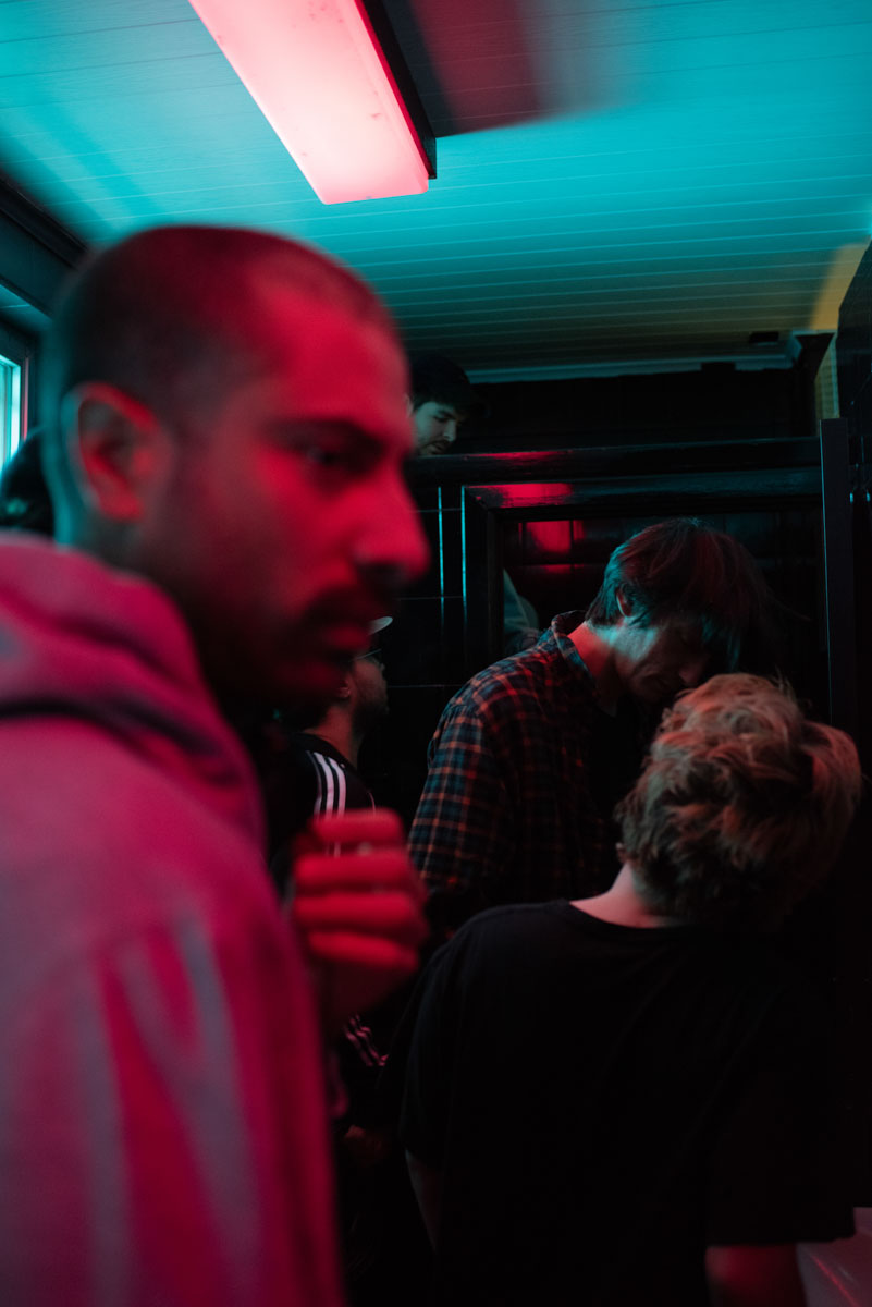 A man in profile in the foreground, lit in red; behind him, two people under a blue-lit ceiling.