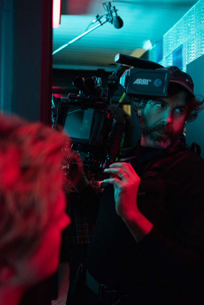 A camera operator holding his camera, lit in red and blue, with a boom microphone overhead.