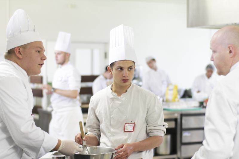 A young cook in a tall white toque holds a whisk over a bowl in a school kitchen, between two chefs.