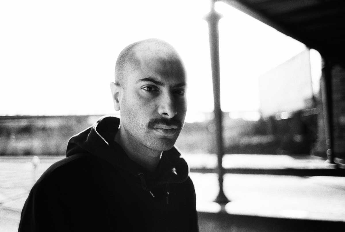 Black-and-white portrait: a man in a dark hooded sweatshirt turned toward the lens, under a platform canopy.