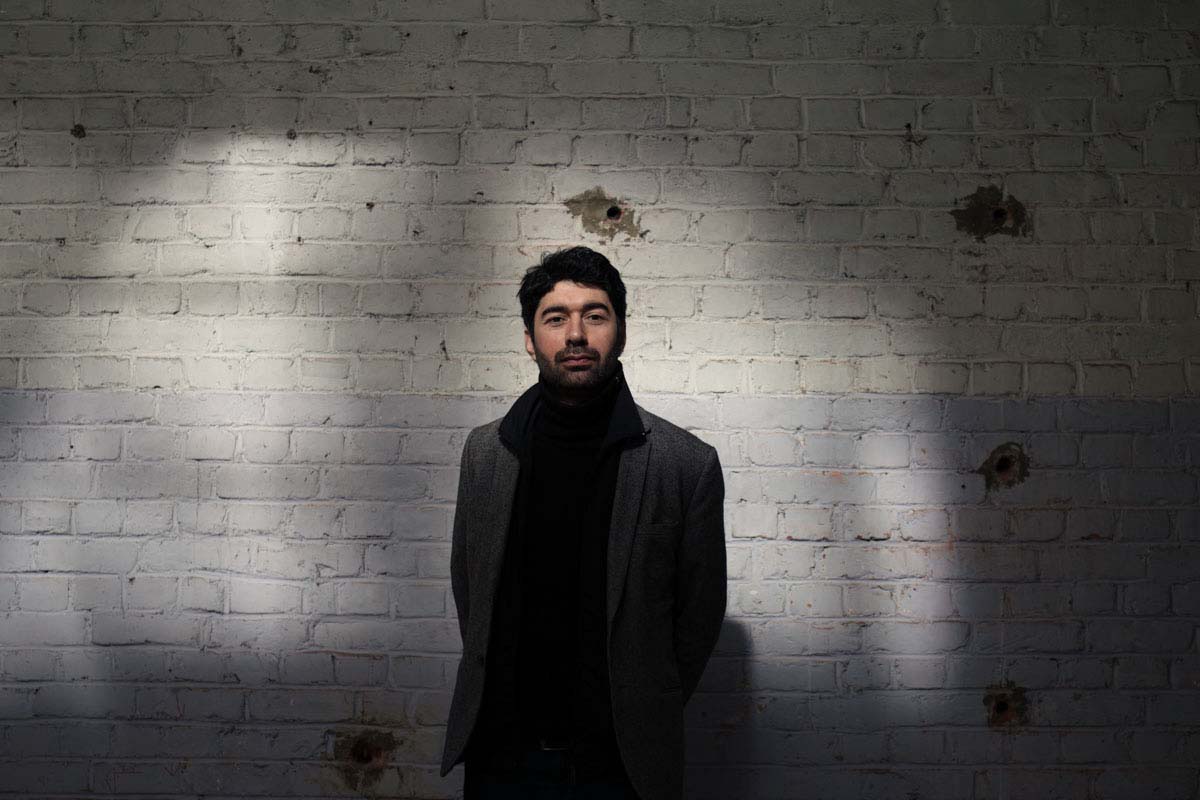 Portrait of a man in a dark jacket standing against a whitewashed brick wall, in a cone of light.